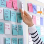 Pessoa interagindo com quadro do método Kanban, movendo post-its coloridos organizados por tarefas e etapas