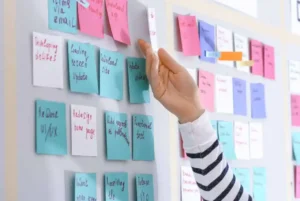 Pessoa interagindo com quadro do método Kanban, movendo post-its coloridos organizados por tarefas e etapas