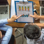 Duas pessoas analisando gráficos impressos e no computador, discutindo resultados e tendências com base em indicadores setoriais.