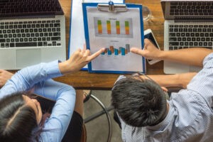 Duas pessoas analisando gráficos impressos e no computador, discutindo resultados e tendências com base em indicadores setoriais.