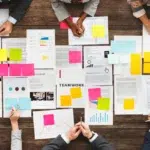 Equipes discutindo documentos de gestão de projetos com post-its, aplicando o PMBOK.