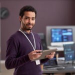 Gestor de automação sorrindo em ambiente de trabalho com computadores e gráficos ao fundo.