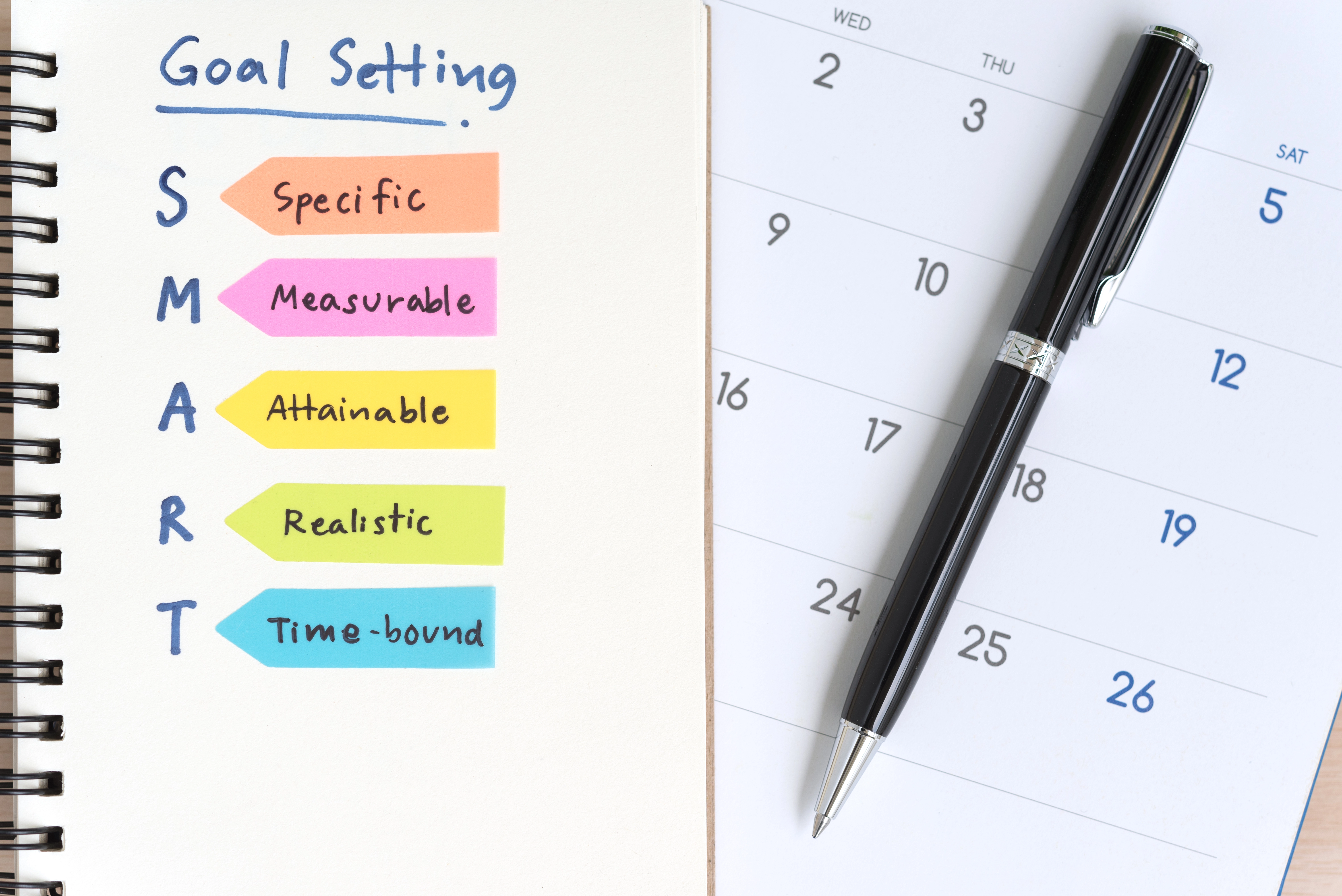 Notebook page with the SMART goal-setting framework written in colorful labels next to a calendar and a black pen, illustrating specific, measurable, attainable, realistic, and time-bound objectives.